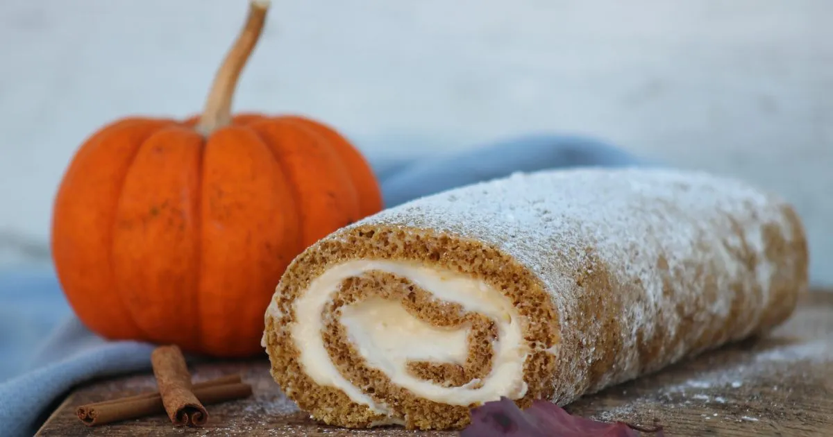 A pumpkin roll cake dusted with powdered sugar, filled with creamy frosting, placed on a wooden board next to a small pumpkin and cinnamon sticks